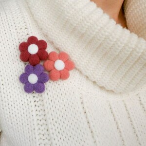 May include: Three felt flower brooches in pink, purple and red with white centres. The brooches are pinned to a white knitted sweater.