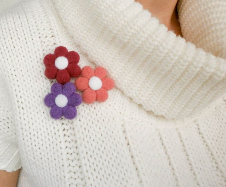 May include: Three felt flower brooches in pink, purple and red with white centres. The brooches are pinned to a white knitted sweater.