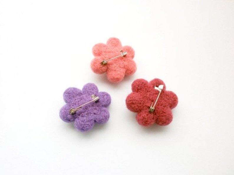 May include: Three felt flower brooches in pink, purple and red with silver safety pins attached to the back.