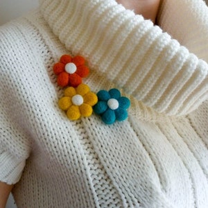 K&ouml;nnte beinhalten: Drei Filzblumenbroschen in Orange, Gelb und Blau. Die Blumen haben wei&szlig;e Zentren und sind in einer Reihe auf einem wei&szlig;en Strickpullover angeordnet.