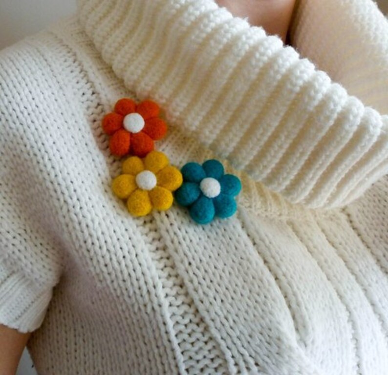 K&ouml;nnte beinhalten: Drei Filzblumenbroschen in Orange, Gelb und Blau. Die Blumen haben wei&szlig;e Zentren und sind in einer Reihe auf einem wei&szlig;en Strickpullover angeordnet.