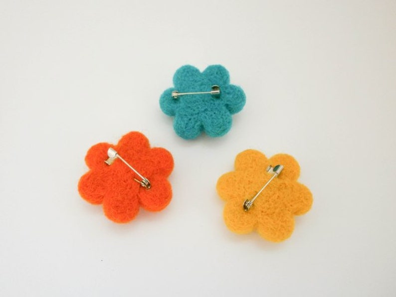 K&ouml;nnte beinhalten: Drei Filzblumenbroschen in T&uuml;rkis, Orange und Gelb. Jede Blume hat eine Sicherheitsnadel auf der R&uuml;ckseite.