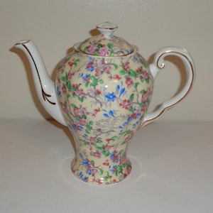 May include: A vintage teapot with a floral pattern in pink, blue, and green on a cream background. The teapot has a white spout and handle with gold trim, and a matching lid with a small knob. A classic piece for tea parties.