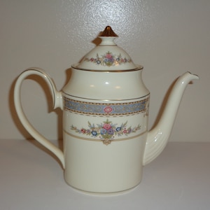 May include: Cream porcelain coffee pot with a lid and a curved handle. The pot has a decorative band with blue, pink, and gold floral patterns. The lid is topped with a gold finial, and gold trim accents the edges.