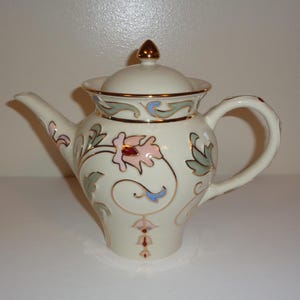 May include: Cream ceramic teapot with gold-coloured detailing and a finial. The teapot is decorated with pink, blue, and green floral designs. The handle and spout are curved. A decorative item for the kitchen or dining room.