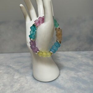 Puede incluir: Un brazalete de cuentas de colores con cuentas de color azul pastel, amarillo, rosa y beige. Las cuentas son facetadas y tienen un acabado mate. El brazalete está enhebrado en un alambre plateado.