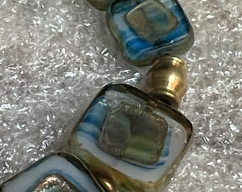 May include: A close-up of a necklace with square-shaped glass beads. The beads are a mix of blue, green, and white with gold accents. The necklace is on a white background.