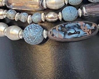 May include: A multi-strand necklace with blue, silver, and brown beads. The beads are various shapes and sizes, including round, oval, and rectangular. The necklace is on a black background.