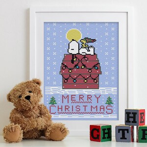 May include: A framed cross-stitch design featuring Snoopy and Woodstock on top of his doghouse decorated with Christmas lights. The text "Merry Christmas" is below the doghouse. The image is displayed on a white shelf with a brown teddy bear and colourful alphabet blocks.