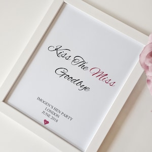May include: White framed print with the text "Kiss The Miss Goodbye" in black cursive script. The word "Miss" is in pink. Below the text is "Imogen's Hen Party, London, June 2018" in black text. A small pink heart is below the text.