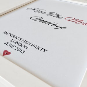 May include: White framed sign with the text "Kiss The Miss Goodbye" in black cursive script. Below the text is "Imogen's Hen Party, London, June 2018" in black print. A small red heart is in the bottom left corner.