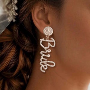 May include: A pair of dangle earrings with the word "Bride" spelled out in rhinestones. The earrings are gold-toned and have a small round rhinestone cluster at the top.
