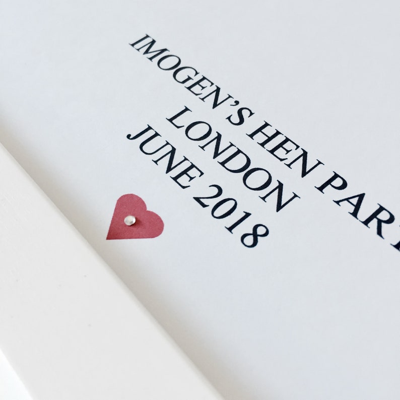 May include: A white card with black text that reads "Imogen's Hen Party London June 2018". A small red heart with a white dot in the center is in the lower left corner of the card.