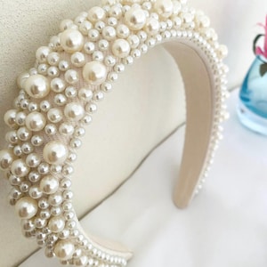 May include: Ivory-colored headband densely adorned with faux pearls of varying sizes. The pearls are clustered together, creating a textured, opulent appearance. The headband's base is a soft, off-white fabric, providing a neutral backdrop for the lustrous pearls. A stylish hair accessory.