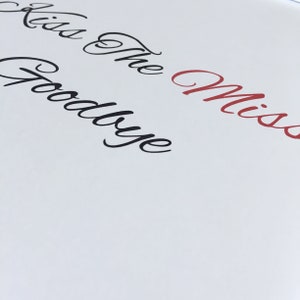 May include: White card with the text "Kiss the Goodbye Miss" written in black and red cursive.