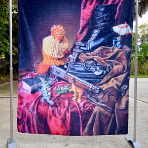 Puede incluir: Una pintura de bodegón con un bolso de cuero marrón, un rifle, un libro, un gorro de punto, una bota, un vaso de líquido y una sombrilla de cóctel. La pintura está sobre un fondo de tela roja y negra.