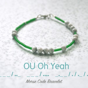 Puede incluir: Un brazalete de cuentas verdes y blancas con un cierre plateado. El brazalete deletrea "OU Oh Yeah" en código Morse.