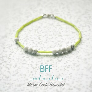 May include: A green and white beaded bracelet with the letters "BFF" and the Morse code for "BFF" written below. The bracelet is on a white background with a speckled pattern.