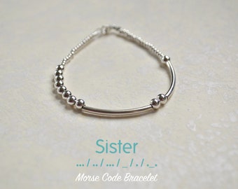 May include: Silver bracelet with a bar and beads spelling out the word "Sister" in Morse code. The bracelet is on a white background.