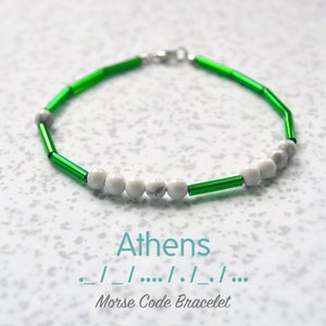 May include: A beaded bracelet with green and white beads. The bracelet features green, cylindrical beads and white, round beads. The word "Athens" and Morse code are printed on the image. The bracelet is on a white speckled surface.