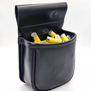 May include: A black leather cartridge bag, open to reveal yellow shotgun shells. The bag has a flap closure and a stitched design. The bag is designed to hold ammunition for hunting or shooting sports.