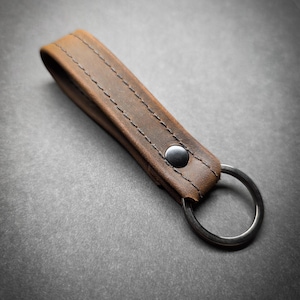 May include: A brown leather keychain with a silver key ring. The leather strap is stitched with dark thread and secured with a black snap. The keychain is designed for holding keys.
