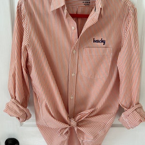 May include: A red and white striped long-sleeved button-down shirt with the text "The Classic Shirt" and "Old Navy" on the label. The shirt has a pocket on the left side with the word "beachy" embroidered on it.
