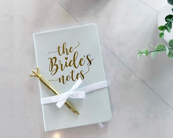 May include: A light blue notebook with a gold inscription that reads "the Bride's notes". A gold pen with a diamond-shaped top is tied to the notebook with a white ribbon.