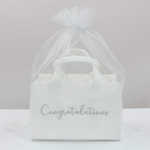 May include: White gift bag with a silver glitter inscription that reads "Congratulations". The bag is made of fabric and has a handle. It is packaged in a white tulle bag.