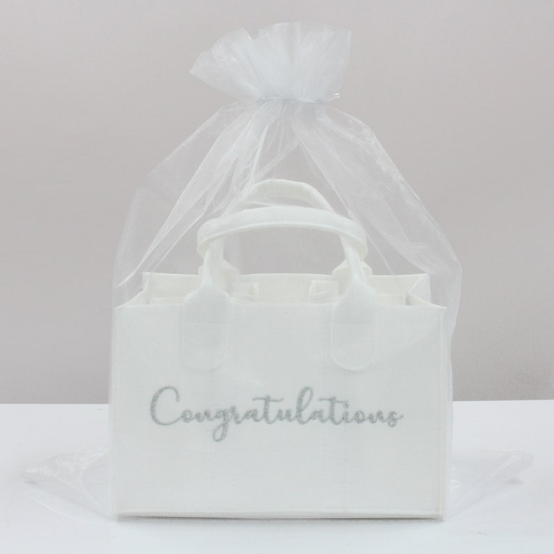 May include: White gift bag with a silver glitter inscription that reads "Congratulations". The bag is made of fabric and has a handle. It is packaged in a white tulle bag.