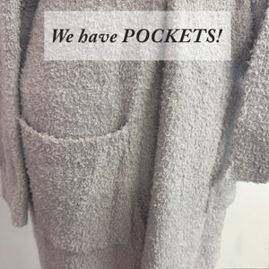 May include: A light gray, fuzzy cardigan with a large pocket is shown. The text "We have POCKETS!" is displayed in a white rectangle across the top of the image. The cardigan has a soft, textured appearance.
