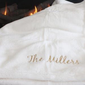 May include: A white, plush blanket with the embroidered text "The Millers" in gold script. The blanket is folded, with a soft, textured appearance. The background shows a fireplace with flames.