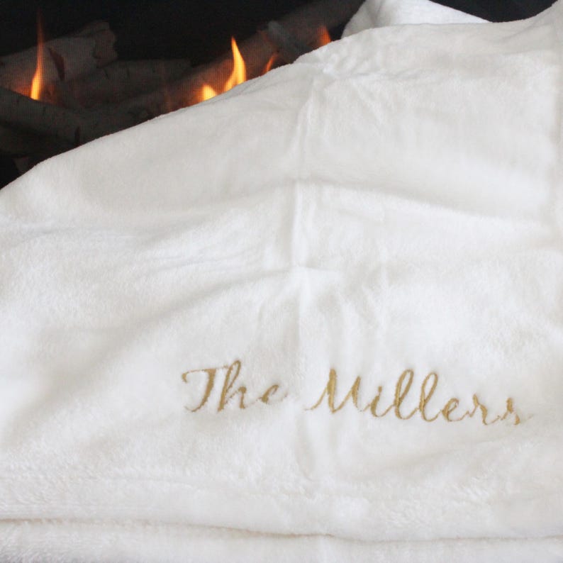 May include: A white, plush blanket with the embroidered text "The Millers" in gold script. The blanket is folded, with a soft, textured appearance. The background shows a fireplace with flames.