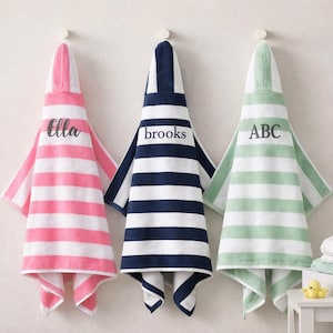 May include: Three hooded baby towels hanging on a wall. The towels are striped in pink and white, navy and white, and green and white. Each towel has a name or initials printed on the hood: "Ella", "brooks", and "ABC".