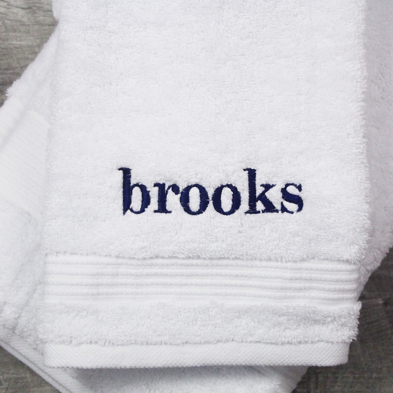 May include: A white terry cloth towel featuring the name brooks embroidered in dark blue lowercase letters. The towel has a textured surface with a horizontal ribbed border detail above the hemline, showcasing a clean and simple design.