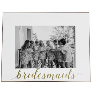 May include: White picture frame with a gold border and the word "bridesmaids" written in gold script. The frame holds a black and white photo of a group of people smiling and holding up glasses.