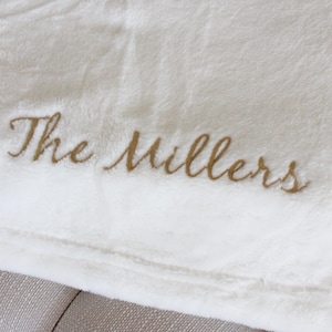 May include: Cream-colored plush blanket with the embroidered text "The Millers" in a gold script. The blanket is folded, showing a soft, textured surface. Ideal for home decor or gifting.