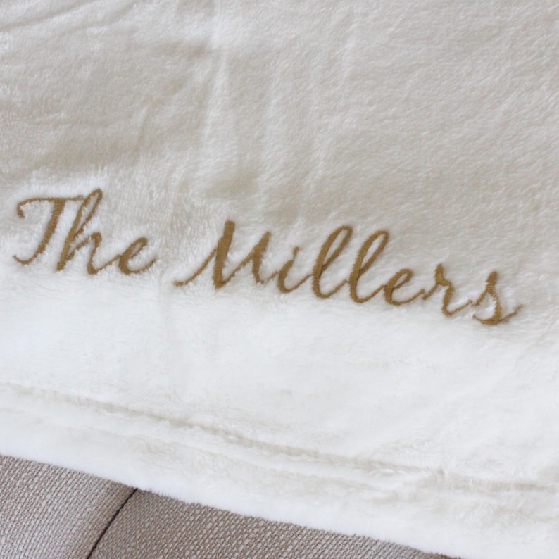 May include: Cream-colored plush blanket with the embroidered text "The Millers" in a gold script. The blanket is folded, showing a soft, textured surface. Ideal for home decor or gifting.