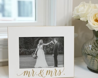 May include: A white picture frame with a gold border and the words "mr. & mrs." in gold script. The frame holds a black and white photo of a wedding couple. A vase of white roses sits nearby.