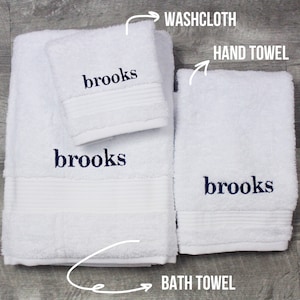 May include: A set of white terry cloth towels including a washcloth, a hand towel, and a bath towel. Each piece features the name brooks embroidered in dark blue thread. The set includes labels pointing to each item with the text WASHCLOTH, HAND TOWEL, and BATH TOWEL.
