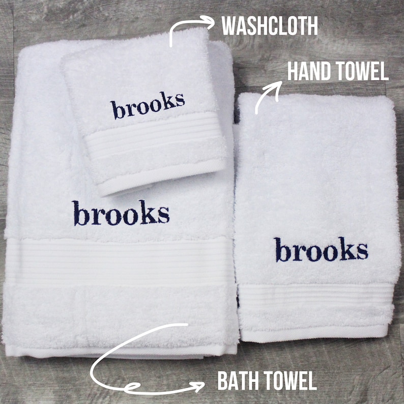 May include: A set of white terry cloth towels including a washcloth, a hand towel, and a bath towel. Each piece features the name brooks embroidered in dark blue thread. The set includes labels pointing to each item with the text WASHCLOTH, HAND TOWEL, and BATH TOWEL.