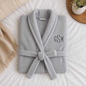 May include: A folded, light gray plush robe with a shawl collar and attached belt. The robe features embroidered initials "GSM" on the chest. The robe is lying on a white bedspread, with a beige blanket and a small plant in the background.