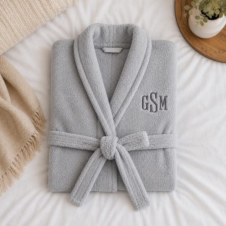 May include: A folded, light gray plush robe with a shawl collar and attached belt. The robe features embroidered initials "GSM" on the chest. The robe is lying on a white bedspread, with a beige blanket and a small plant in the background.