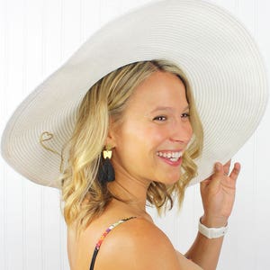May include: A white wide-brimmed sun hat with a gold heart detail. The hat is worn by a person with blonde hair and black tassel earrings.