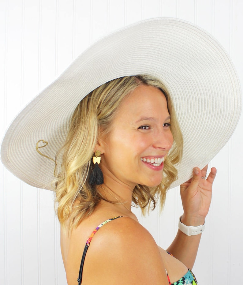 May include: A white wide-brimmed sun hat with a gold heart detail. The hat is worn by a person with blonde hair and black tassel earrings.