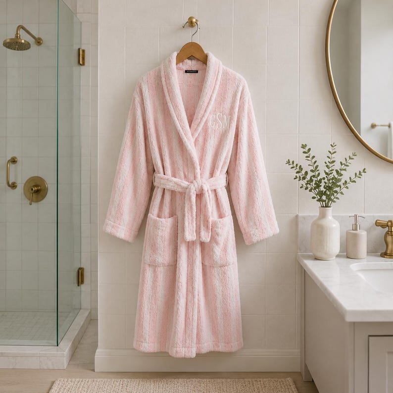 May include: A pink and white striped plush robe with a matching belt and pockets hangs on a wooden hanger. The robe has the initials "CSM" embroidered on the chest. The robe is displayed in a bathroom setting.