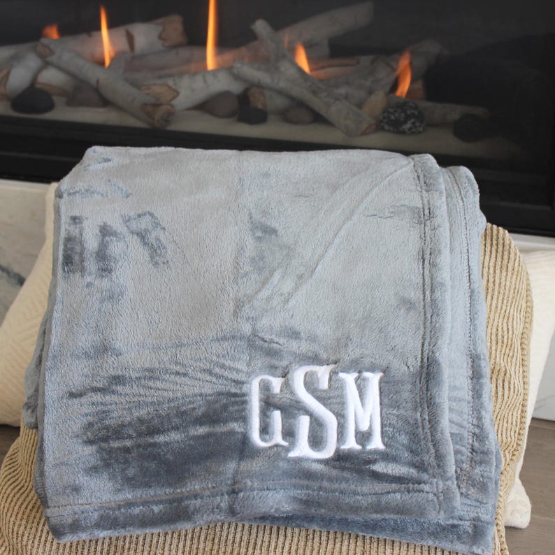 May include: A soft, gray blanket with white embroidered initials "GSM". The blanket is folded and resting on a striped cushion, with a fireplace and burning logs in the background. The blanket appears plush and cozy.