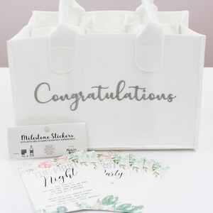 May include: White gift bag with the word "Congratulations" embroidered in silver. The bag has six pockets for bottles. The bag is accompanied by a set of milestone stickers and cards with floral designs and text that reads "The First Night" and "First Party".