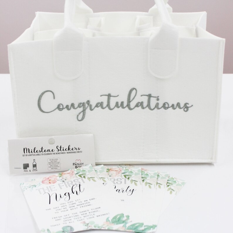 May include: White gift bag with the word "Congratulations" embroidered in silver. The bag has six pockets for bottles. The bag is accompanied by a set of milestone stickers and cards with floral designs and text that reads "The First Night" and "First Party".