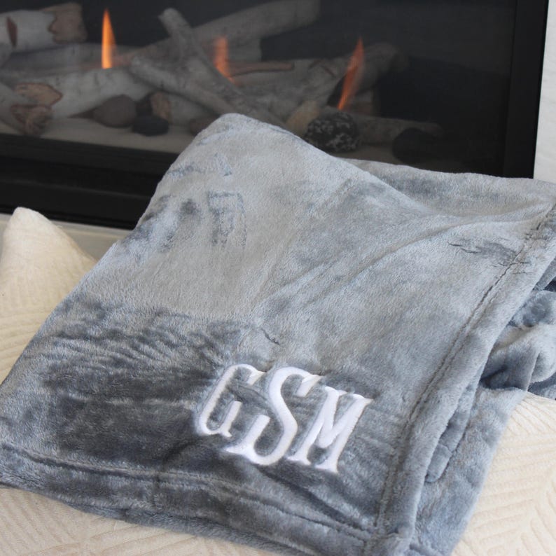 May include: A soft, gray plush blanket with white embroidered initials "GSM". The blanket is folded and resting on a cream-colored surface, with a fireplace and burning flames in the background. The blanket appears to be made of a soft, velvety material.